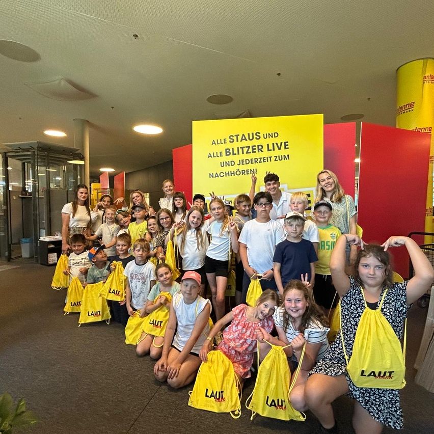 Eine Gruppe von Kindern mit gelben Taschen und einem gelben Schild, auf dem 'Alle Staus und alle Blitzer live und jederzeit zum Nachhören!' steht, posiert für ein Foto.