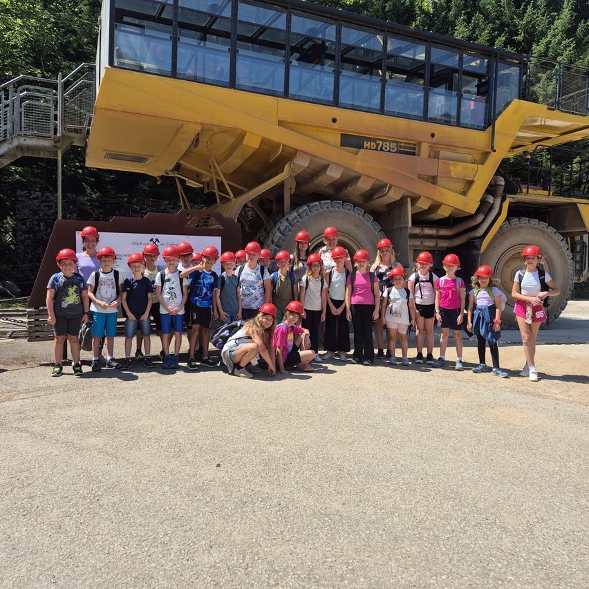 Eine Gruppe von Kindern und Erwachsenen in roten Helmen posiert vor einem großen gelben Bergbaulastwagen.