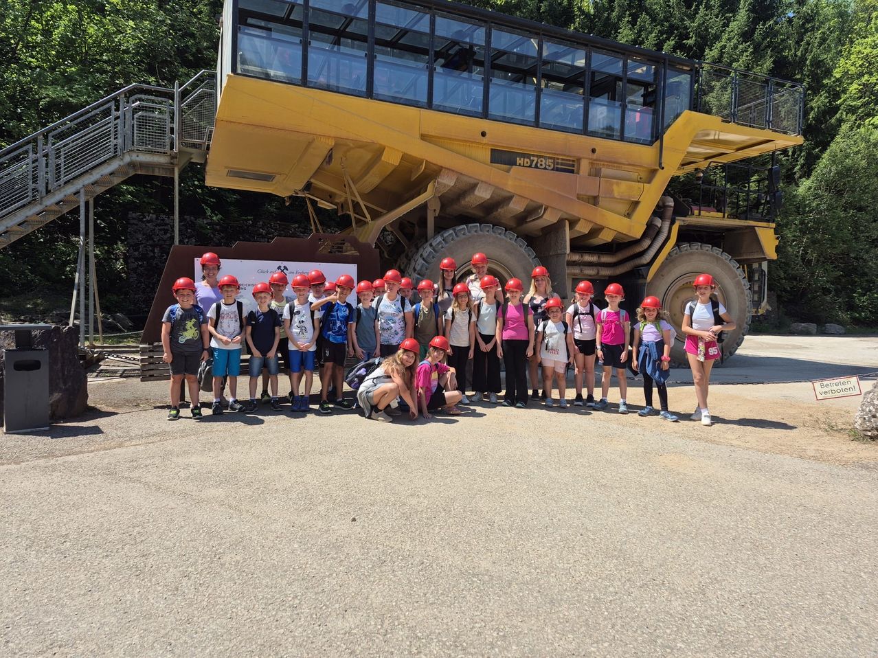 Eine Gruppe von Kindern und Erwachsenen in roten Helmen posiert vor einem großen gelben Bergbaulastwagen.