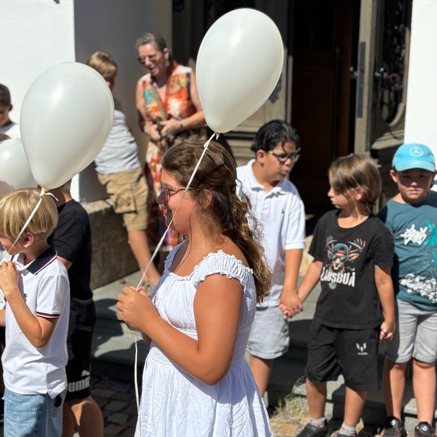 Eine Gruppe von Kindern, die weiße Luftballons halten, steht vor einem Gebäude, mit einer Frau und anderen Kindern im Hintergrund.