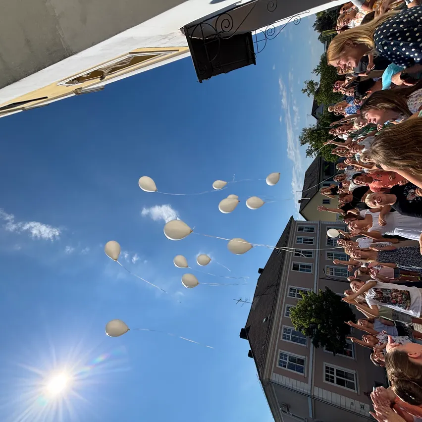 Eine Menschenmenge versammelt sich im Freien und lässt weiße Luftballons in einen klaren blauen Himmel aufsteigen. Die Ballons schweben nach oben, einige sind bereits außer Sichtweite.