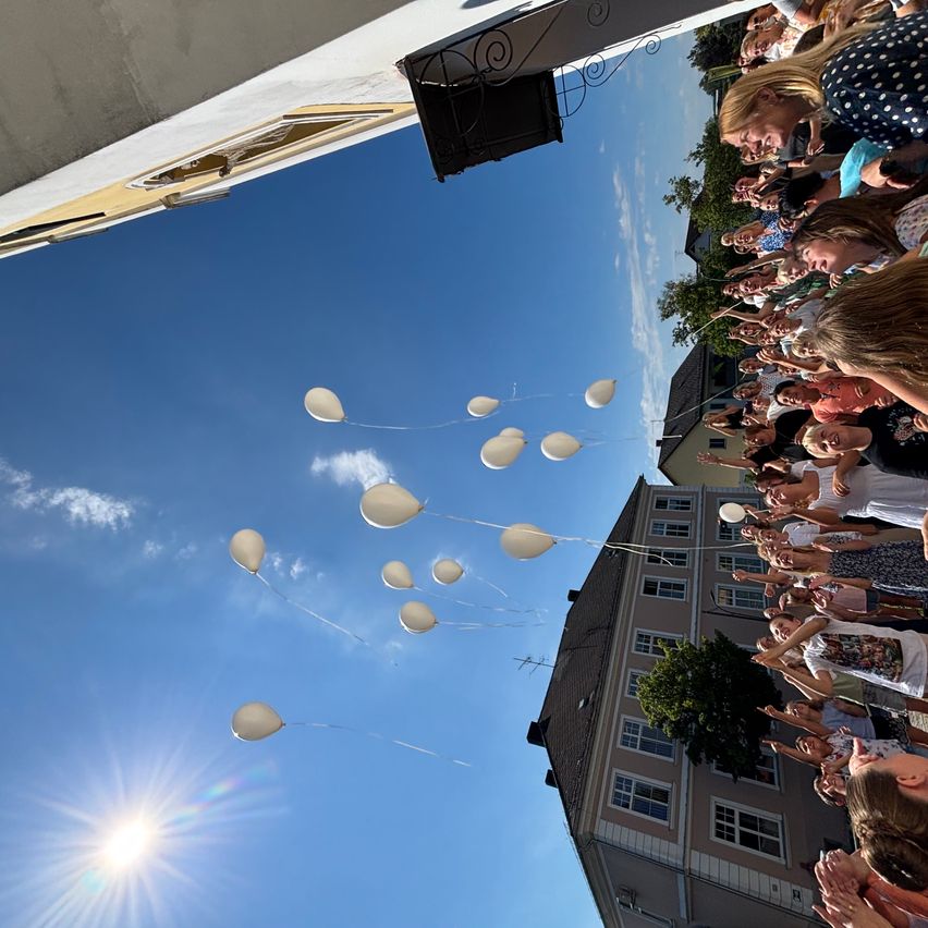 Eine Menschenmenge versammelt sich im Freien und lässt weiße Luftballons in einen klaren blauen Himmel aufsteigen. Die Ballons schweben nach oben, einige sind bereits außer Sichtweite.
