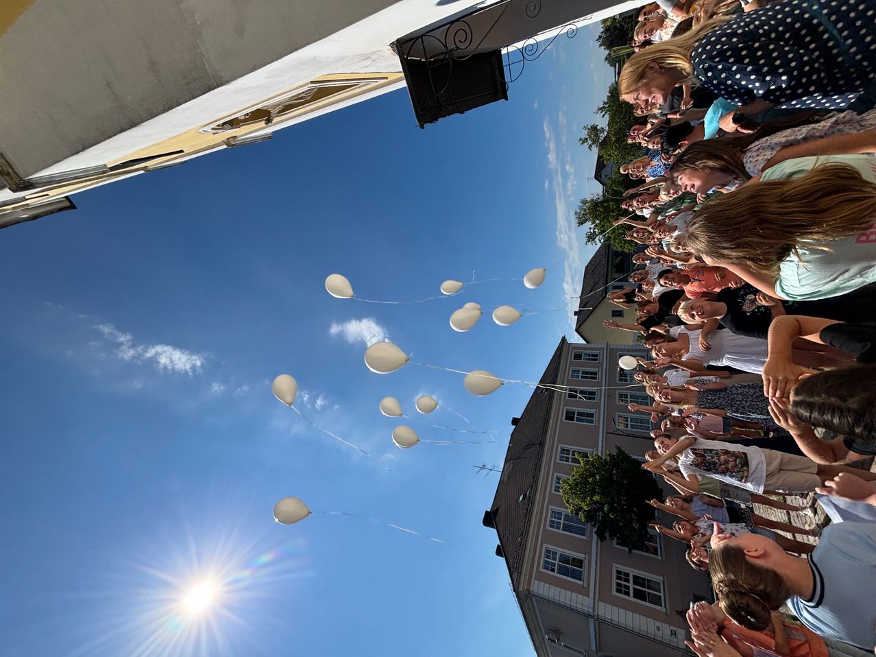 Eine Menschenmenge versammelt sich im Freien und lässt weiße Luftballons in einen klaren blauen Himmel aufsteigen. Die Ballons schweben nach oben, einige sind bereits außer Sichtweite.