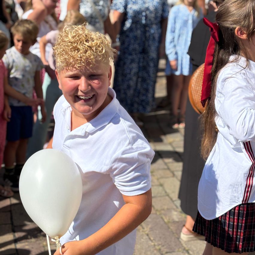 Ein junger Junge mit lockigem blondem Haar hält einen weißen Ballon und lächelt fröhlich, während er unter anderen Kindern in einer überfüllten Außenumgebung steht.