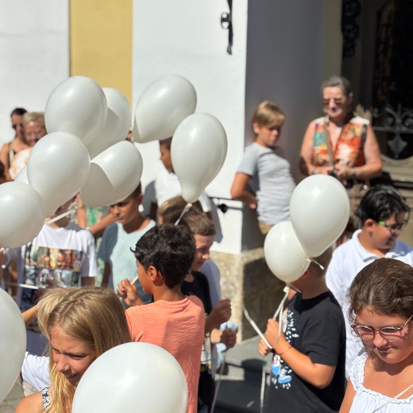 Eine Gruppe von Kindern und Erwachsenen, die weiße Luftballons halten, in einer Außenumgebung, mit einem Gebäude und Personen im Hintergrund.