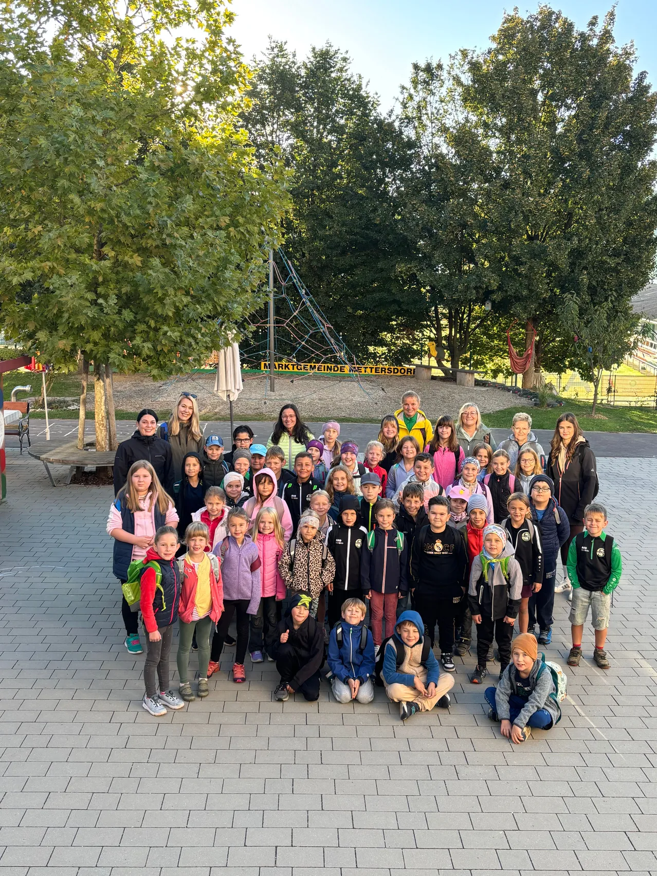 Eine Gruppe von Kindern und Erwachsenen posiert für ein Foto in einem Außenbereich mit Bäumen und einem Spielplatz im Hintergrund.