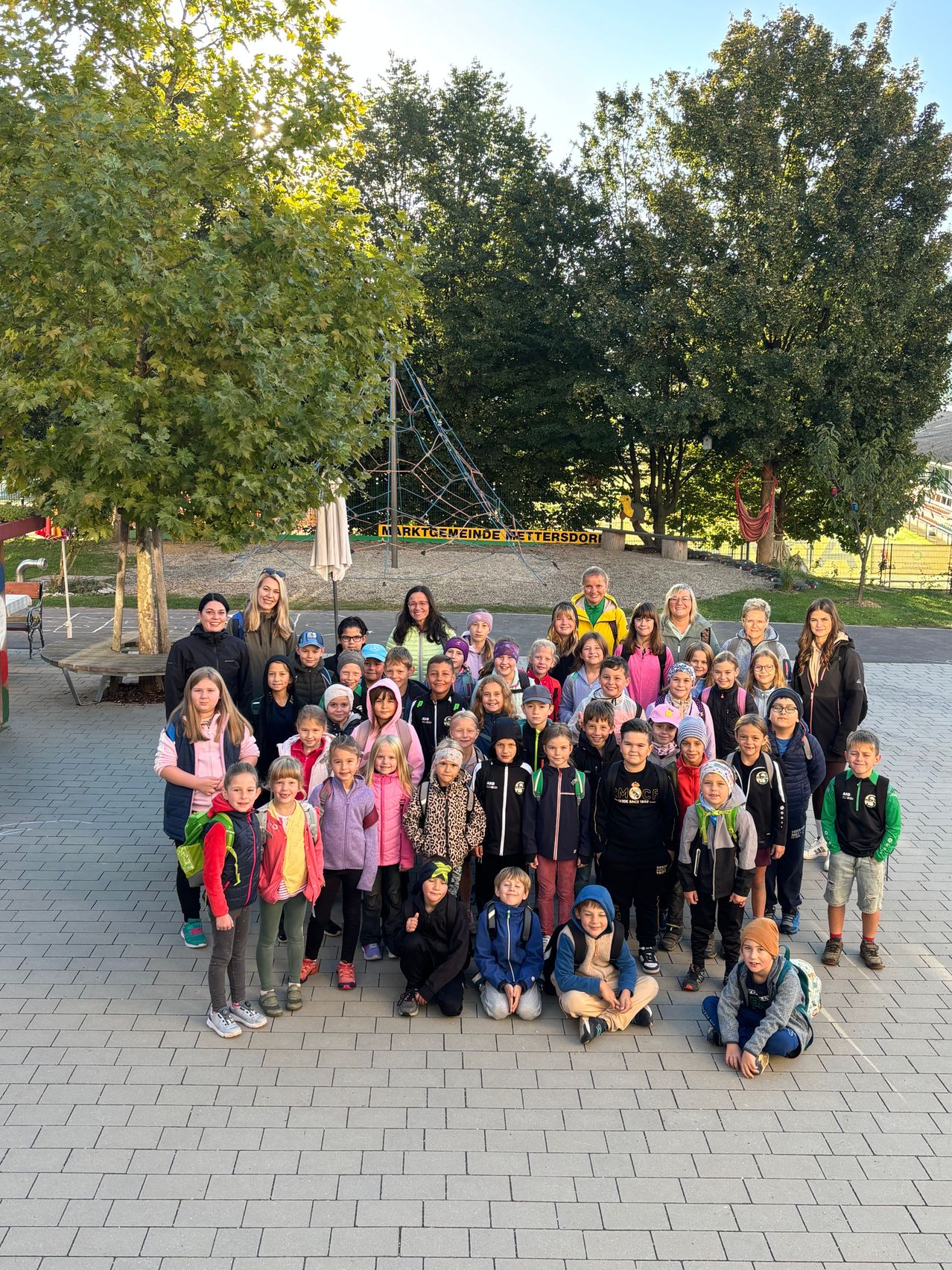 Eine Gruppe von Kindern und Erwachsenen posiert für ein Foto in einem Außenbereich mit Bäumen und einem Spielplatz im Hintergrund.