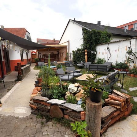 Ein Außenbereich mit einem Ziegelpflanzenbehälter, verschiedenen Pflanzen und Sitzgelegenheiten. Der Patio ist von Gebäuden und Grün umgeben.