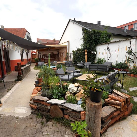 Ein Außenbereich mit einem Ziegelpflanzenbehälter, verschiedenen Pflanzen und Sitzgelegenheiten. Der Patio ist von Gebäuden und Grün umgeben.