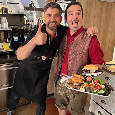 Zwei Männer posieren für ein Bild in der Küche. Einer trägt eine schwarze Schürze und der andere ein rotes Hemd und traditionelle Shorts. Sie halten einen Tablett mit Essen.
