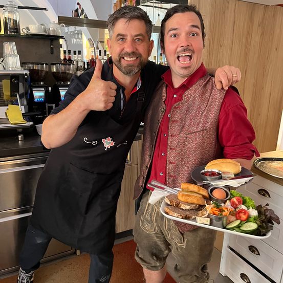 Zwei Männer posieren für ein Bild in der Küche. Einer trägt eine schwarze Schürze und der andere ein rotes Hemd und traditionelle Shorts. Sie halten einen Tablett mit Essen.