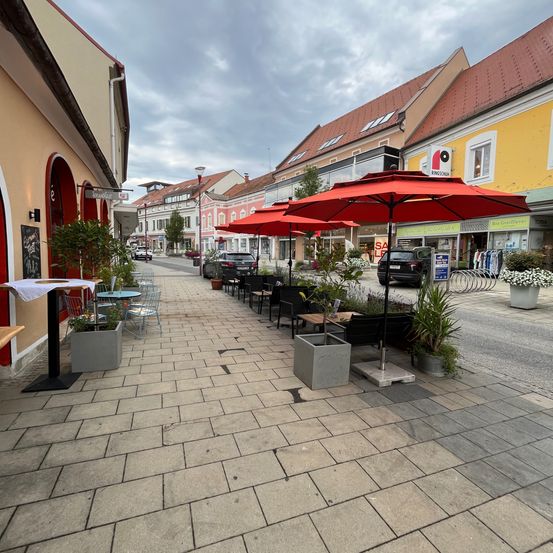 Outdoor-Essensbereich mit roten Sonnenschirmen auf einem Bürgersteig. Gebäude mit Geschäften auf der rechten Seite und geparkte Autos in der Ferne.