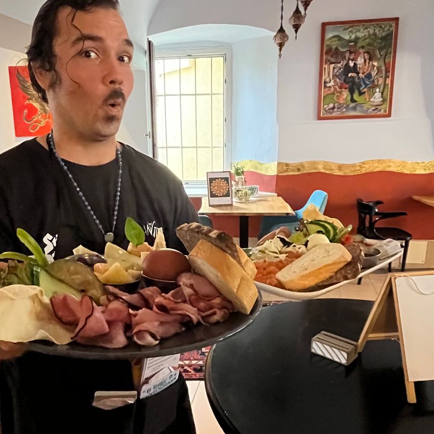 Ein Mann hält eine große Platte mit verschiedenen Speisen, einschließlich Fleisch, Gemüse und Brot, in einem Restaurant.