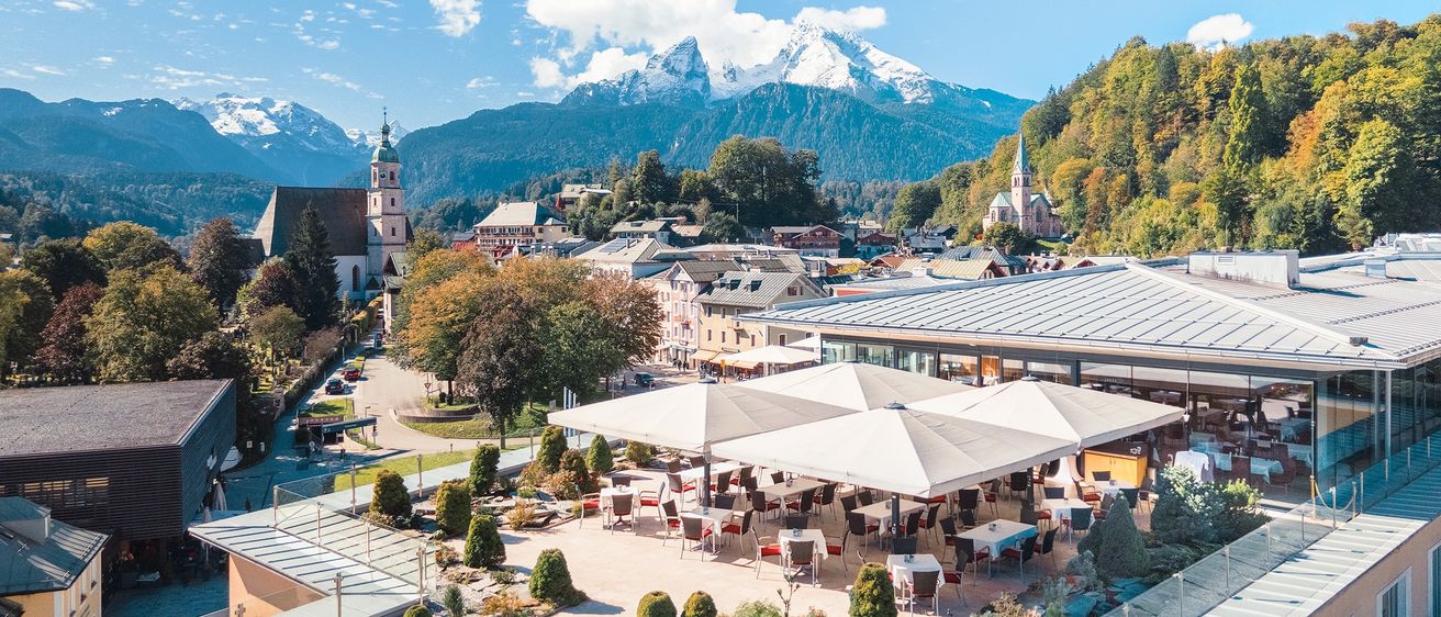 Eine Vogelperspektive auf eine Stadt mit Bergen im Hintergrund. Die Stadt hat Gebäude mit Dächern und eine Kirche. Es gibt Bäume, Pflanzen und eine Straße. Es gibt ein Restaurant mit Tischen und Stühlen unter einem Regenschirm. Es gibt ein Gebäude mit einem Glasdach.