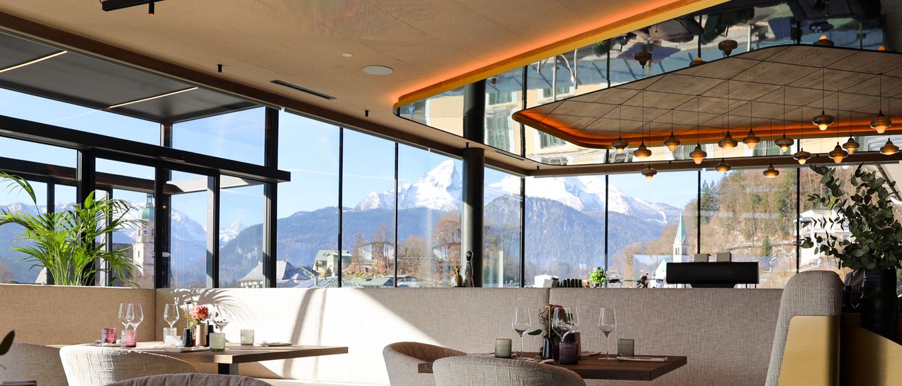 Ein modern gestaltetes Innenrestaurant mit großen Fenstern, die verschneite Berge zeigen. Tische mit Weingläsern und Pflanzen.