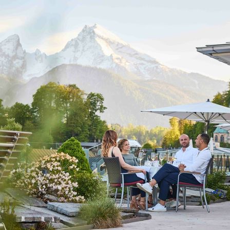 Eine Gruppe von Menschen genießt ein Essen auf einer Terrasse im Freien, mit Bergen und Bäumen im Hintergrund.
