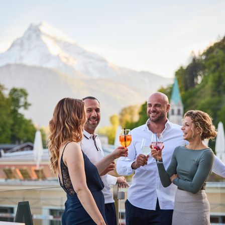 Drei Personen stoßen mit Weingläsern auf einer Terrasse an, im Hintergrund ein Gebirgszug.