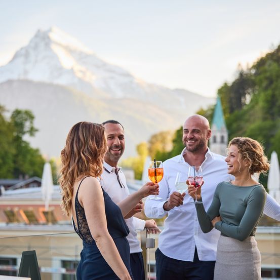 Drei Personen stoßen mit Weingläsern auf einer Terrasse an, im Hintergrund ein Gebirgszug.