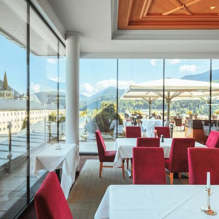 Ein elegantes Restaurant mit großen Glaswänden, das einen Blick auf Berge und ein nahes Gebäude bietet. Rote Stühle umgeben Tische mit weißen Tischdecken. Ein Schirm und eine Kerze auf einem Kerzenständer sind sichtbar.
