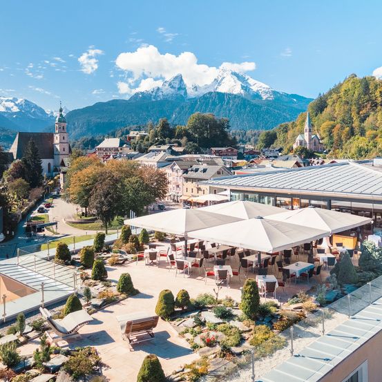 Luftaufnahme einer Stadt mit einem Berg im Hintergrund. Die Stadt hat Gebäude, eine Kirche und einen Garten mit Stühlen und Tischen.