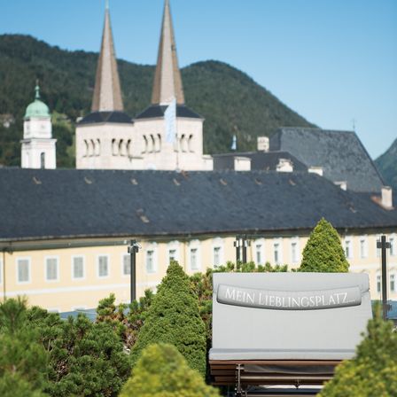 Ein Blick auf ein großes gelbes Gebäude mit einem grauen Dach und Türmen. Im Vordergrund steht ein Lounge-Sessel mit einem Schild, das 'Mein Lieblingsplatz' besagt, umgeben von grünen Büschen. Im Hintergrund sind Berge zu sehen.