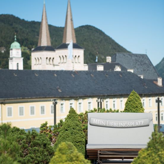 Ein Blick auf ein großes gelbes Gebäude mit einem grauen Dach und Türmen. Im Vordergrund steht ein Lounge-Sessel mit einem Schild, das 'Mein Lieblingsplatz' besagt, umgeben von grünen Büschen. Im Hintergrund sind Berge zu sehen.
