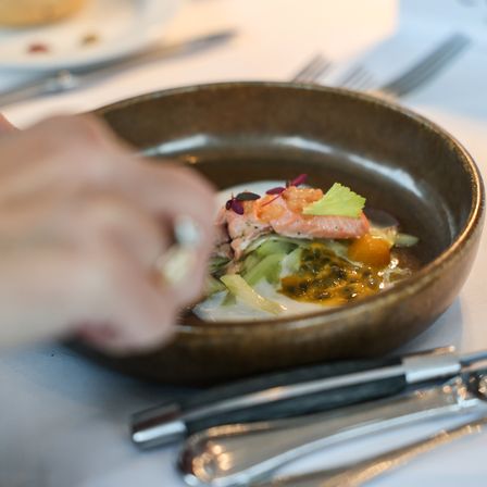 Eine Person garniert eine Schüssel mit Lachs und Zitrone mit Kräutern. Ein Teller, Messer und Gabel sind in der Nähe.