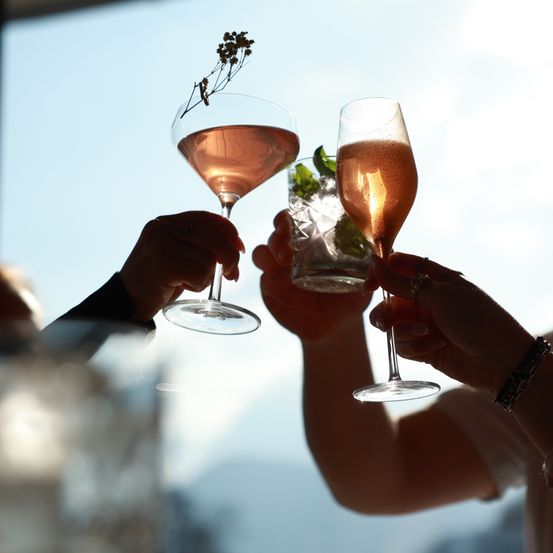 Zwei Hände stoßen mit Gläsern rosa Cocktail und Champagner bei strahlend blauem Himmel an.