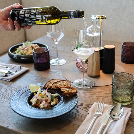 Eine Person gießt eine Flasche Weißwein in ein Weinglas auf einem Holztisch mit Essen und Besteck.