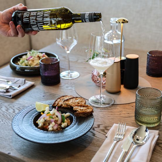 Eine Person gießt eine Flasche Weißwein in ein Weinglas auf einem Holztisch mit Essen und Besteck.