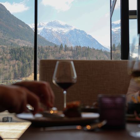 Eine Person isst an einem Tisch mit Blick auf verschneite Berge und grüne Bäume. Ein Weinglas und eine Kerze stehen auf dem Tisch.
