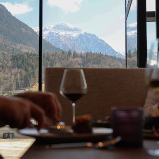 Eine Person isst an einem Tisch mit Blick auf verschneite Berge und grüne Bäume. Ein Weinglas und eine Kerze stehen auf dem Tisch.