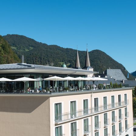 Ein Blick von oben auf ein modernes Hotel mit einer großen Glasterrasse, umgeben von Bergen und Grün. Das Gebäude hat mehrere Stockwerke, mit vielen Fenstern und einigen Personen, die auf der Terrasse sitzen.
