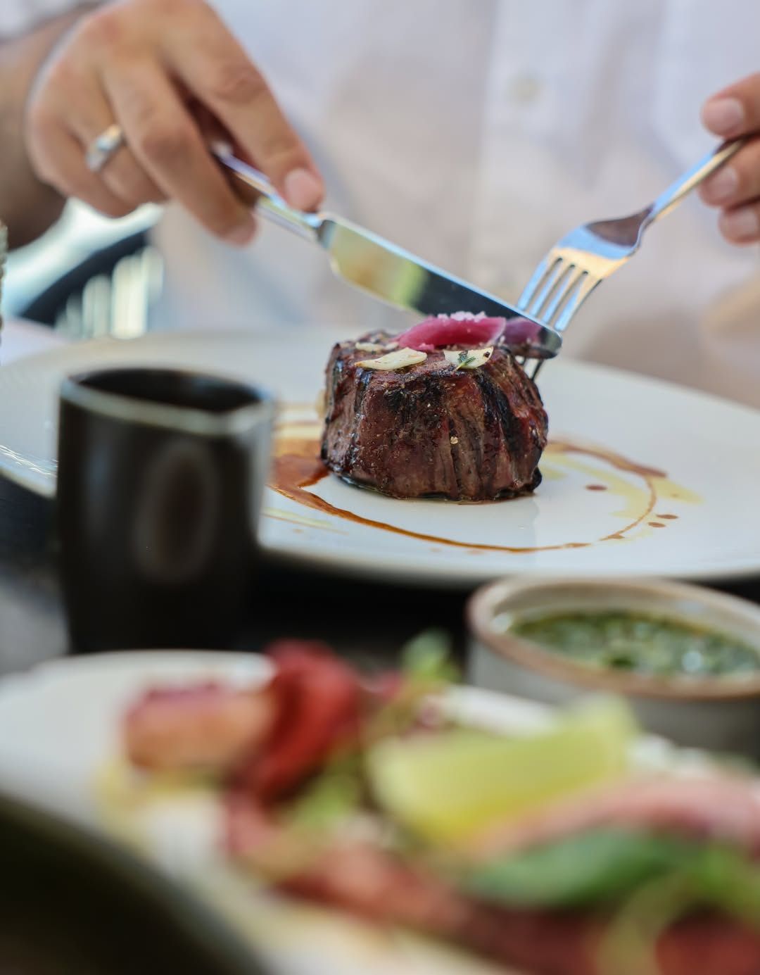 A person is cutting a steak with a knife and fork on a white plate. The steak has a red onion slice and a green herb on top.