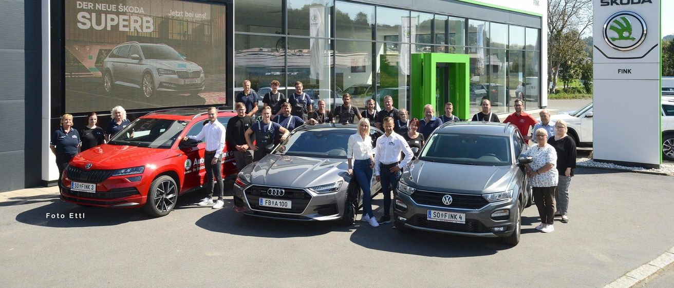 Eine Gruppe von Menschen posiert mit drei Autos vor einem Skoda-Händler. Ein Plakat mit einem Skoda-Auto wird im Schaufenster gezeigt.