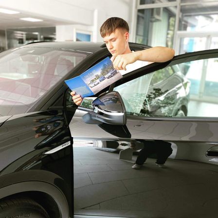Ein junger Mann hält eine Broschüre neben einem schwarzen Auto und betrachtet sie genau in einer Ausstellung.
