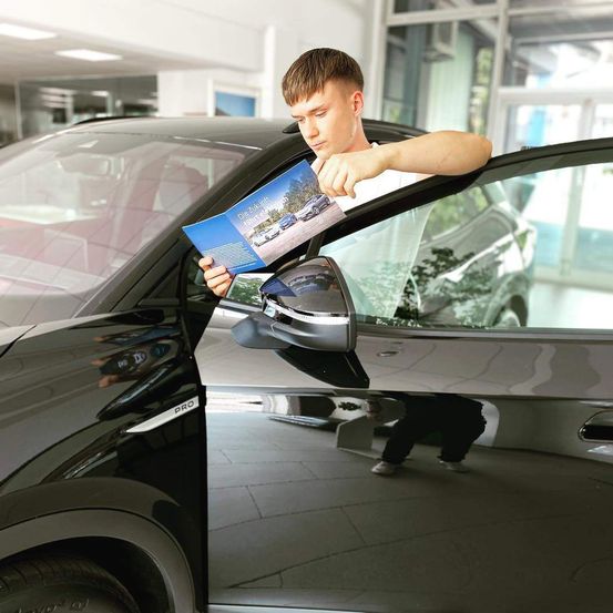 Ein junger Mann hält eine Broschüre neben einem schwarzen Auto und betrachtet sie genau in einer Ausstellung.
