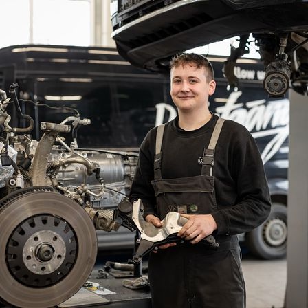 Ein junger Mann steht in einer Garage mit einer Bremsscheibe in den Händen, lächelt und schaut zur Kamera. Hinter ihm steht ein Auto auf der Hebebühne.