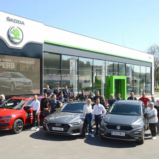 Gruppe von Menschen posiert vor einem Skoda-Händler mit verschiedenen Autos. Das Gebäude hat ein großes Skoda-Logo. Die Autos umfassen einen roten Skoda, einen grauen Audi und einen grauen Volkswagen.
