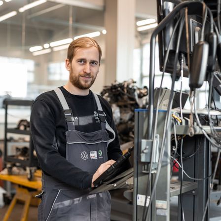 Ein Mann in einer Autowerkstatt, der eine Schürze trägt, lächelt und hält ein Tablet. Hinter ihm stehen Maschinen und Geräte, und die Decke ist beleuchtet.