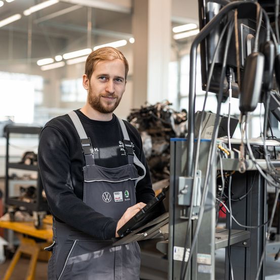 Ein Mann in einer Autowerkstatt, der eine Schürze trägt, lächelt und hält ein Tablet. Hinter ihm stehen Maschinen und Geräte, und die Decke ist beleuchtet.