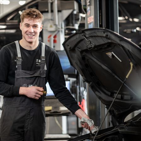 Ein Mann in einer Volkswagen-Garage steht neben einem Auto mit offenem Motorhaube und lächelt. Er hält ein Tuch in der rechten Hand.