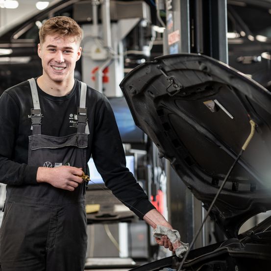 Ein Mann in einer Volkswagen-Garage steht neben einem Auto mit offenem Motorhaube und lächelt. Er hält ein Tuch in der rechten Hand.