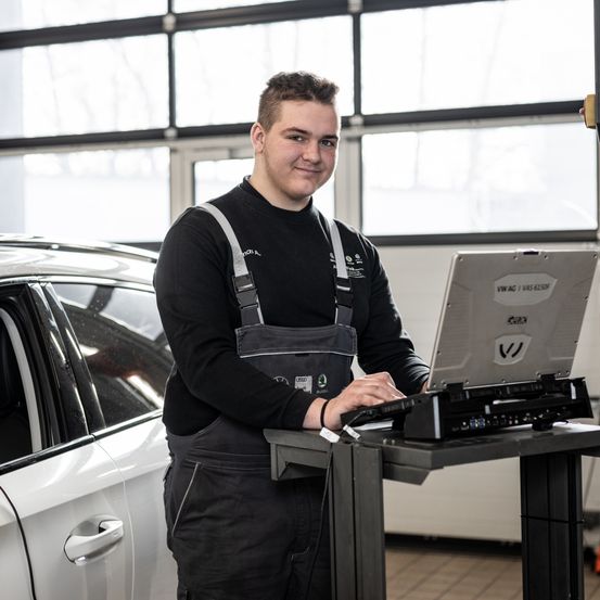 Ein Mann steht in einer Autowerkstatt und arbeitet an einem Laptop auf einem Ständer vor einem weißen Auto. Er trägt ein schwarzes Shirt und eine Schürze.