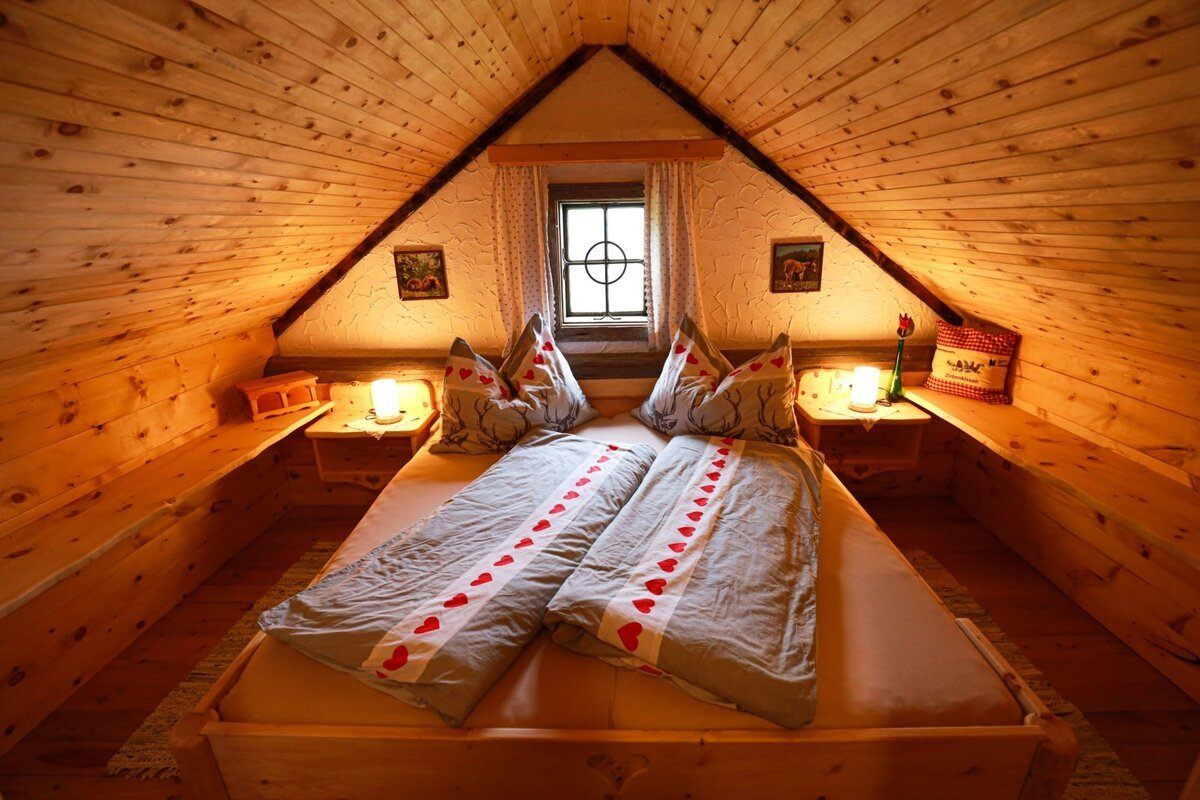 A cozy attic bedroom with wooden ceiling and walls, featuring two twin beds adorned with heart-patterned blankets. Nightstands with lamps and picture frames flank the beds. A window with curtains adds natural light.