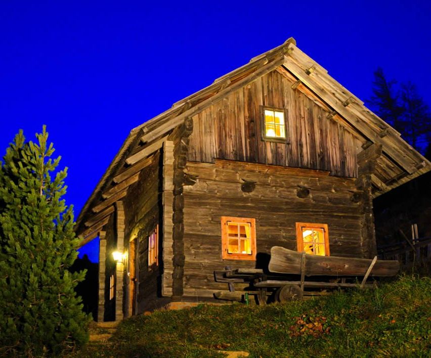 A log cabin is illuminated at night with a blue sky above and windows glowing. There is a bench and a tree in front of the cabin.