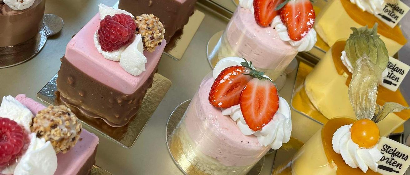 Eine Reihe von Mini-Desserts mit verschiedenen Toppings wie Erdbeeren, Schlagsahne und Nüssen. Jedes Dessert ist auf einem goldenen Ständer ausgestellt. Einige sind schokoladig, andere sind rosa und wieder andere haben Früchte darauf.