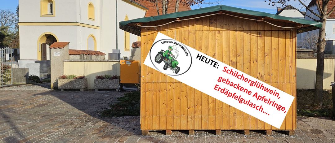 Ein Holzschuppen mit grünem Dach zeigt ein Schild mit der Werbung für Kartoffeln heute. Ein Traktorlogo und der Text befinden sich in einem Kreis.