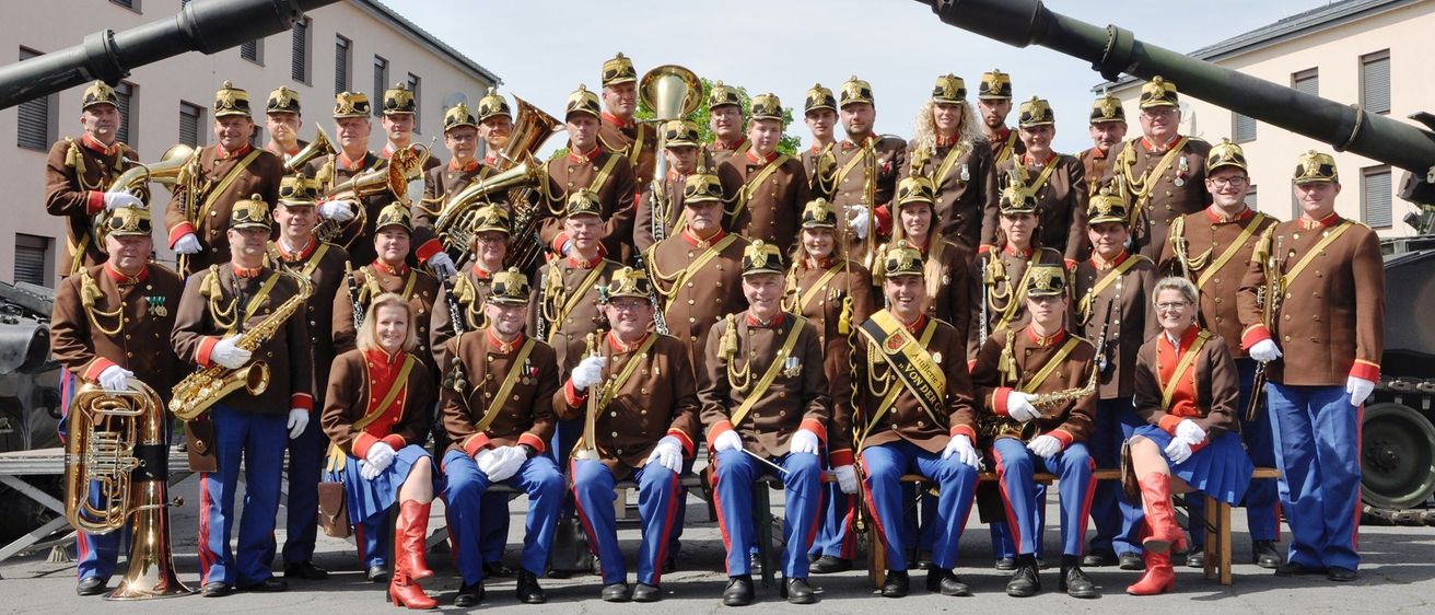 Eine Militärkapelle posiert vor zwei großen Kanonen, einige Mitglieder halten Instrumente und tragen Uniformen mit Medaillen und Schärpen.