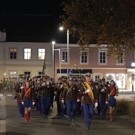 Ein Militärorchester marschiert am Abend, gekleidet in Uniformen und mit Instrumenten, vor einem Gebäude mit mehreren Fenstern.